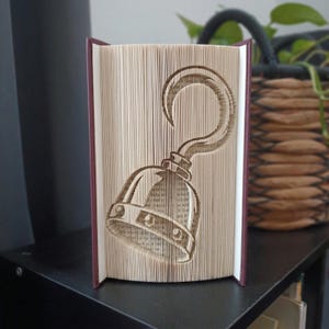 Peut inclure: Une sculpture de livre avec des pages pliées formant la silhouette distincte d'un crochet de pirate courbé. Les bords du papier sont pliés avec précision pour créer la forme du crochet, avec les couvertures du livre visibles sur les côtés.