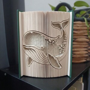Peut inclure: Une sculpture de livre avec des pages pliées pour former la silhouette tridimensionnelle d'une baleine. Le livre possède une couverture vert foncé et les pages sont de couleur crème avec du texte imprimé noir visible sur le motif.