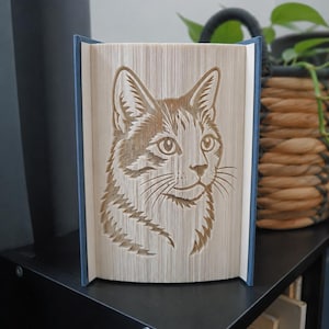 Può includere: Una scultura in libro piegato che presenta un design dettagliato del muso di un gatto, creato piegando con precisione le pagine di un libro con copertina rigida. Il libro ha una copertina blu e le pagine formano la sagoma del gatto.