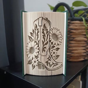 May include: A folded book art sculpture featuring a cowboy boot design surrounded by sunflowers. The pages are precisely creased to create the three-dimensional image of the boot and floral elements, with a dark green hardback cover visible.