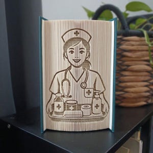 Op de afbeelding: Een gevouwen boekkunstsculptuur van een verpleegkundige met een kapje en uniform, inclusief een stethoscoop om de nek. De verpleegkundige houdt een dienblad vast met medische benodigdheden zoals flesjes, een potje en een rol tape, gevormd uit pagina's.