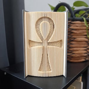 May include: A folded book sculpture featuring an ankh symbol created by precisely folding the individual pages of a hardback book. The cream-coloured paper edges form the distinct cross shape with a loop at the top, centred within the book.