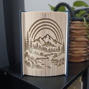 May include: A folded book art sculpture featuring a mountain range landscape with a sun or rainbow arch above, pine trees on the sides, and a river flowing through the foreground, all created by precisely folding the pages of a hardback book.