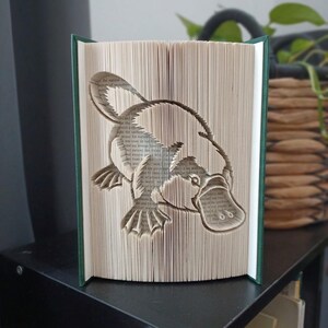 May include: A folded book art sculpture featuring a detailed platypus silhouette created by precisely folding the individual pages of a hardback book. The book has dark green covers and the pages show visible printed text throughout the design.