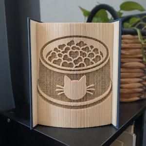 Può includere: Una scultura di libro creata piegando le pagine per formare un'immagine tridimensionale di una ciotola per animali piena di croccantini a forma di cuore. La parte anteriore della ciotola presenta una sagoma di testa di gatto con baffi.