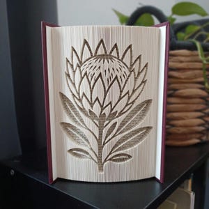 Op de afbeelding: Een boeksculptuur met een gevouwen paginadesign dat de vorm van een proteabloem creëert. Het boek heeft een donkerrode kaft en de pagina's zijn wit met zichtbare zwarte tekst, zorgvuldig gevouwen om het gedetailleerde bloemmotief te vormen.