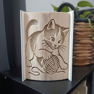 Peut inclure: Une sculpture en livre plié représentant un chaton jouant avec une pelote de laine. Les pages du livre sont pliées avec précision pour créer l'image tridimensionnelle du chat, avec le texte des pages originales visible sur l'ensemble.