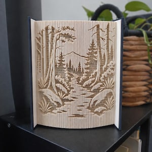 May include: A folded book art sculpture featuring a detailed landscape scene with a central path leading towards a mountain range, surrounded by tall pine trees and low-lying foliage, all created by precisely folding the pages of a hardback book.