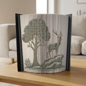 May include: A decorative book sculpture featuring a tree and deer design. The pages are folded to create the image, with a black frame. The artwork is in shades of green and gray, and sits on a wooden table.