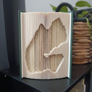 Peut inclure: Une sculpture de livre avec des pages pliées formant la lettre G. Le livre possède une couverture vert foncé et les pages intérieures de couleur crème sont minutieusement pliées et courbées pour créer la forme de la lettre G majuscule.