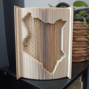 May include: A book sculpture featuring folded pages that form the distinct outline of the map of Kenya. The cream-colored paper edges are precisely creased to create a three-dimensional silhouette of the country, revealing printed text.