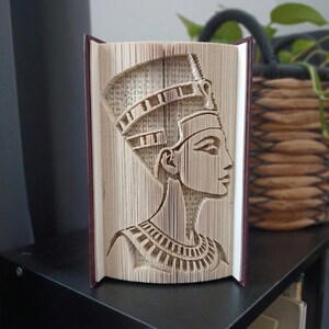 Op de afbeelding: Een boeksculptuur met gevouwen paginakunst van de Egyptische koningin Nefertiti in profiel. De pagina's zijn nauwkeurig gevouwen om de vorm van de kroon, gelaatstrekken en de versierde kraag te vormen, geplaatst in een donkerrode boekomslag.