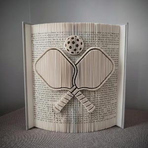 Peut inclure: Une sculpture d'art de livre unique en son genre, sur le thème du pickleball. Le livre ouvert présente une balle et deux raquettes de pickleball, découpées avec précision dans les pages. Les tons neutres du papier créent un effet visuel saisissant, idéal pour les amateurs de sport.