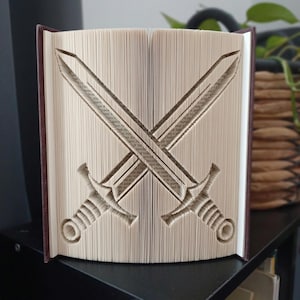 May include: A book sculpture featuring folded pages that form the shape of two crossed swords. The book has a dark brown cover and the pages are cream-coloured, with the sword design clearly defined by the precise folding of each individual page.
