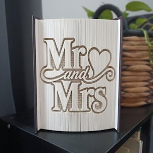 Op de afbeelding: Een gevouwen boekkunstsculptuur met de tekst Mr and Mrs en een hartsymbool. De pagina's zijn nauwkeurig gevouwen om de driedimensionale letters en de hartvorm te creëren, waarbij de donkere boekomslagen aan de zijkanten zichtbaar zijn.