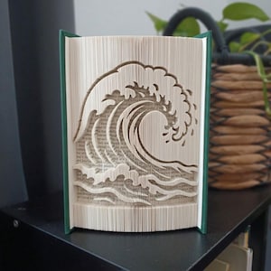 May include: A book sculpture featuring folded pages that form the shape of a large crashing ocean wave. The book has a dark green cover and the interior pages are cream-colored with visible printed text throughout the folded paper design.