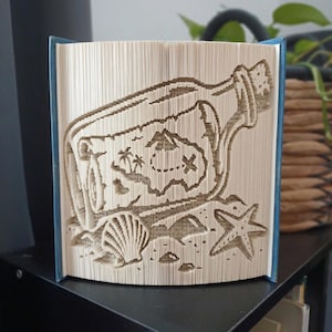 May include: A folded book art sculpture featuring a message in a bottle design. The pages are intricately folded to reveal a silhouette of a glass bottle containing a treasure map with palm trees and an X mark, accompanied by a seashell and starfish.