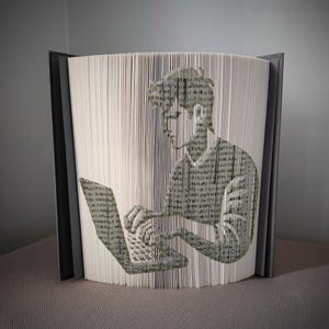 Pode incluir: Escultura de arte de livro única que retrata uma pessoa usando um laptop. A imagem da pessoa e do laptop é criada cortando e dobrando com precisão as páginas de um livro. O livro tem uma capa cinza.