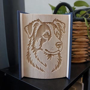 Op de afbeelding: Een boeksculptuur met een gevouwen pagina-ontwerp in de vorm van een hondenkop. De pagina's zijn nauwkeurig gevouwen om het silhouet van een hondengezicht te creëren met gedetailleerde vachtstructuren, ogen en een neus op een donkerblauwe kaft.