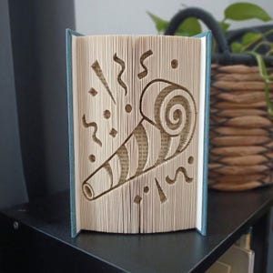 Peut inclure: Une sculpture en livre plié représentant un sifflet de fête, créée en pliant avec précision les pages d'un livre à couverture rigide. Les bords des pages couleur crème forment la silhouette d'une sarbacane entourée de confettis.