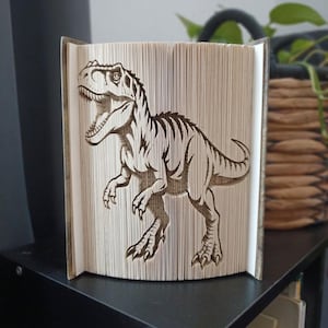 May include: A folded book art sculpture featuring a detailed silhouette of a Tyrannosaurus Rex dinosaur. The pages of the book are precisely folded to create the three-dimensional image of the creature, showing its open mouth, sharp teeth, and tail.