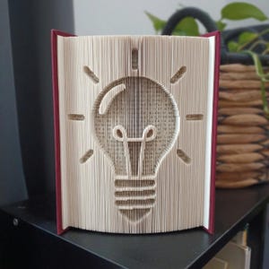 May include: A folded book art sculpture featuring a light bulb design created by precisely folding the pages of a book. The book has a dark red cover and the pages are arranged to form the shape of a glowing light bulb with rays.
