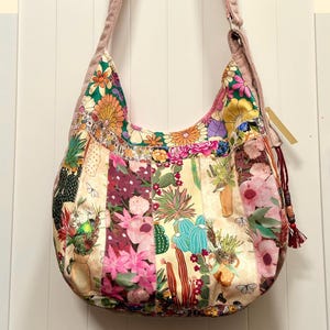 May include: A hobo-style shoulder bag featuring a patchwork design of various floral and cactus prints in shades of pink, green, orange, and yellow. The bag has a wide, solid dusty pink shoulder strap and a decorative tassel with wooden beads.