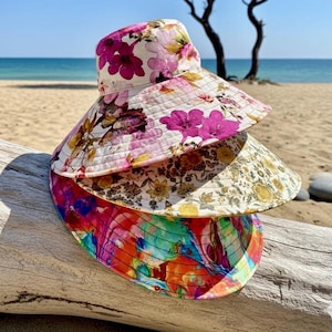 May include: Three stacked bucket hats featuring distinct patterns. The top hat has a white base with large pink and purple floral prints. The middle hat displays a dense yellow and brown botanical design. The bottom hat features a vibrant multicoloured abstract pattern.