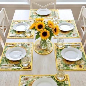 May include: A dining table set for six with sunflower-themed placemats, coasters, and a centerpiece. Each place setting includes a white plate, cutlery, and a glass of water. The placemats and coasters feature a yellow and green floral pattern with sunflowers.