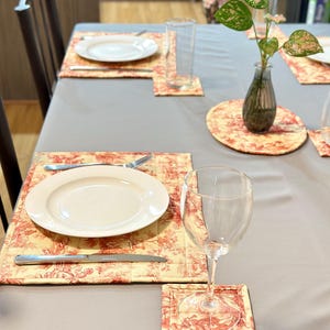 May include: A dining table set with a grey tablecloth, featuring place settings with white plates, silverware, and matching placemats and coasters. The placemats and coasters have a red and cream toile pattern. A clear wine glass and a vase with greenery are also on the table.