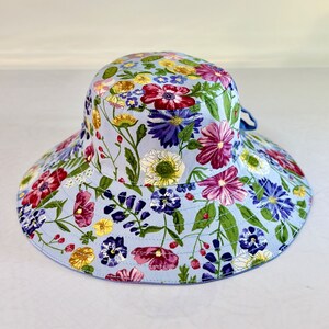 May include: A light blue sun hat with a floral print featuring pink, purple, yellow, and green flowers. The hat has a wide brim and a tied bow.