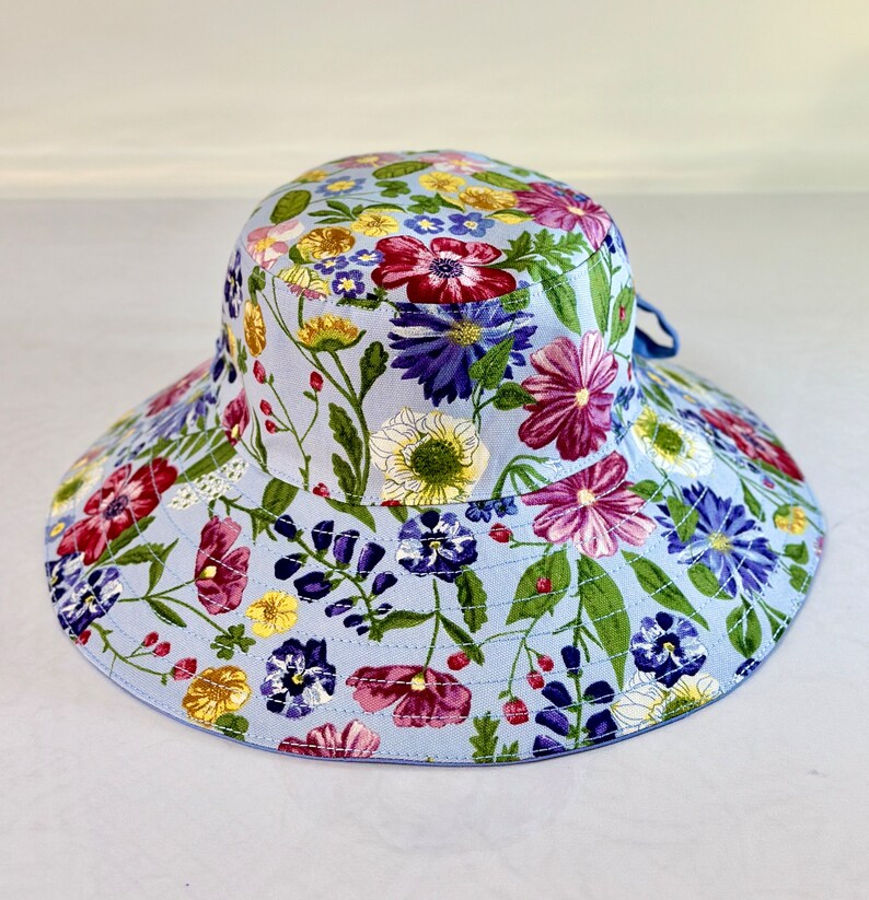 May include: A light blue sun hat with a floral print featuring pink, purple, yellow, and green flowers. The hat has a wide brim and a tied bow.