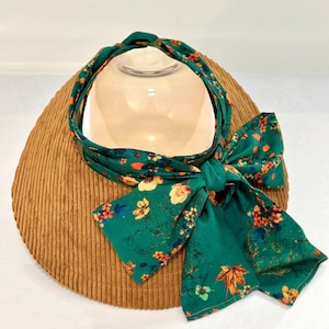 Puede incluir: Una diadema verde con un estampado floral y un lazo. La diadema está envuelta alrededor de un sombrero marrón con textura de pana. El estampado floral incluye flores y hojas naranjas, amarillas y azules.