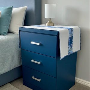 May include: A navy blue three-drawer bedside table with silver handles. A white and blue patterned table runner is on top, with a small white lamp. The bedside table is next to a bed with blue pillows.