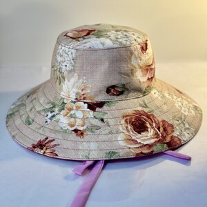 May include: A wide-brimmed sun hat with a floral print in shades of pink, brown, and white. The hat has a light brown fabric base and a purple ribbon tied at the back.