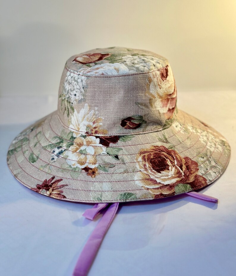 May include: A wide-brimmed sun hat with a floral print in shades of pink, brown, and white. The hat has a light brown fabric base and a purple ribbon tied at the back.