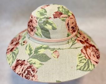 Puede incluir: Un sombrero de sol de ala ancha con un patrón floral. El sombrero está hecho de una tela tejida y tiene una cinta rosa alrededor de la corona.