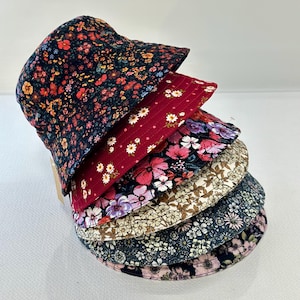 May include: A stack of six bucket hats featuring various floral patterns. The hats display diverse colour palettes including dark navy with orange flowers, deep red with white blossoms, pink and purple florals, and beige with brown botanical prints.