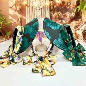 May include: Two sun visors with a dark green floral pattern. The visors have a wide brim and a ribbon tie. The ribbons are a mix of yellow, orange, and blue floral patterns. The visors are on a white surface.