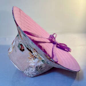 May include: A pink and floral sun hat with a purple ribbon tied around the crown. The hat is made of a lightweight fabric and has a wide brim.