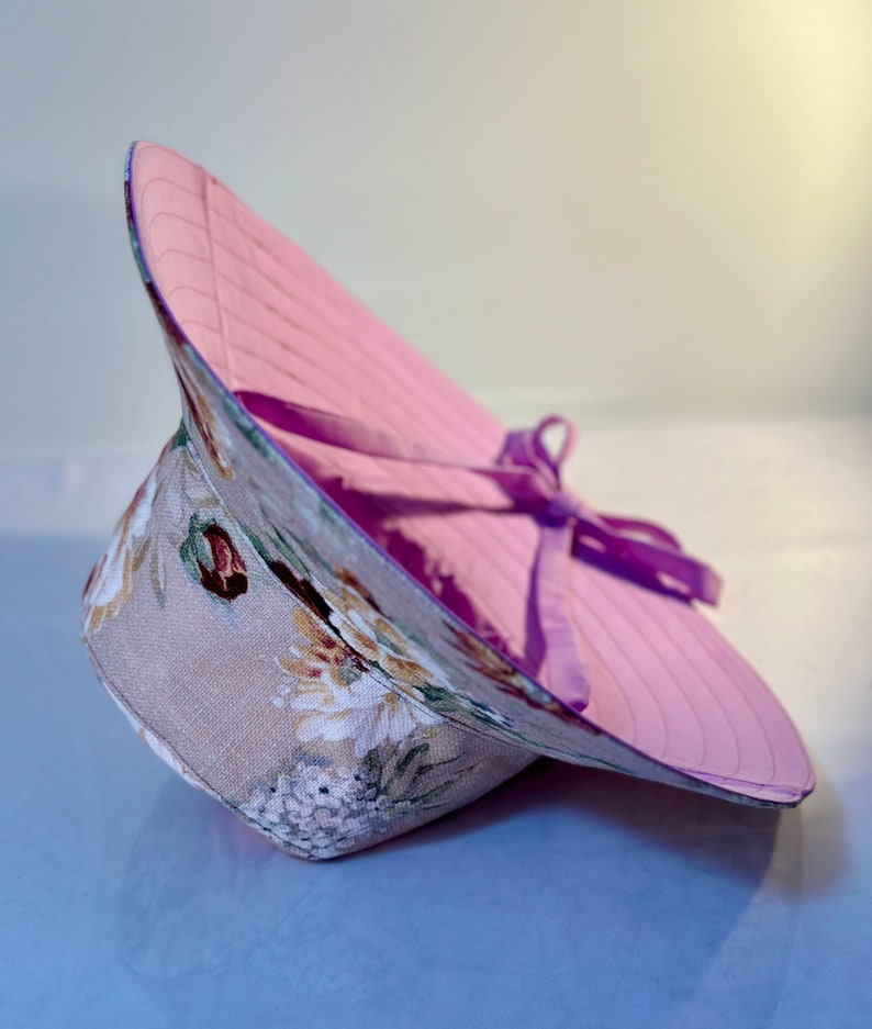 May include: A pink and floral sun hat with a purple ribbon tied around the crown. The hat is made of a lightweight fabric and has a wide brim.