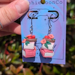 May include: Dangle earrings featuring miniature stacks of books in shades of pink and white, topped with small red cherries and pink flowers. The charms are crafted from clay and hang from silver-toned metal hooks on a purple display card.
