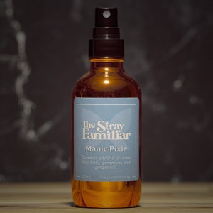 May include: A brown glass bottle with a black spray nozzle and a blue label that reads "the Stray Familiar Manic Pixie Contains a blend of cedar, bay, basil, geranium, and ginger oils. 120mL Fragranted spray"