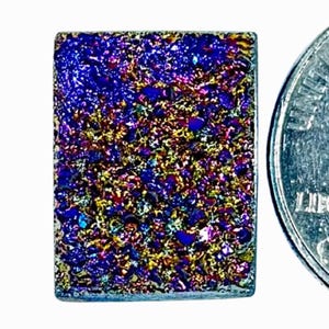 May include: A rectangular dichroic glass piece with a vibrant, multicolored surface. The colors include blues, purples, golds, and greens, creating a shimmering effect. A US quarter coin is next to the glass piece for size comparison.