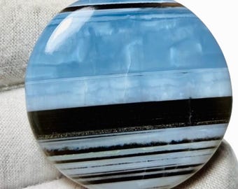 May include: A round, polished gemstone with a striking color pattern. The stone features bands of light blue, black, and white, creating a layered effect. The gemstone is held in a white gloved hand.