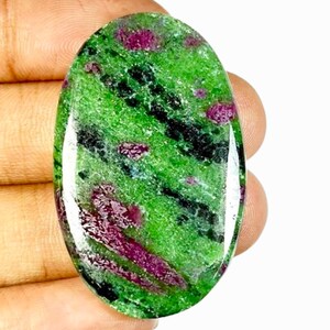 Peut inclure: Pierre précieuse ovale avec une base verte vive, parsemée d'inclusions noires et rouge foncé. La surface polie reflète la lumière, soulignant les motifs et couleurs naturels. Une pierre unique et attrayante.