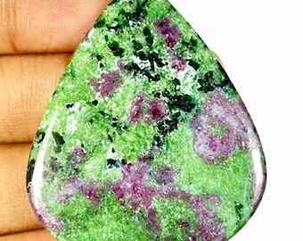 Pode incluir: Uma pedra preciosa em forma de gota com uma superfície polida. A pedra apresenta uma cor de base verde vibrante, salpicada de preto e manchas de vermelho profundo. A pedra preciosa é segurada em uma mão, mostrando sua beleza natural.