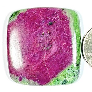 Op de afbeelding: Een vierkant geslepen edelsteen met een levendig magenta centrum en een groene rand. De steen ligt naast een munt ter vergelijking van de grootte. Het oppervlak van de edelsteen heeft natuurlijke patronen en variaties.