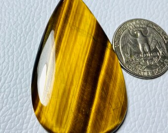 May include: A teardrop-shaped cabochon of tiger's eye gemstone, with a golden brown color and a chatoyant effect. The stone is approximately the size of a US quarter.
