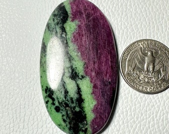 May include: An oval-shaped polished gemstone featuring a vibrant mix of deep magenta, light green, and black mineral inclusions. A United States quarter coin is positioned to the right of the stone to provide a clear sense of scale for the object.